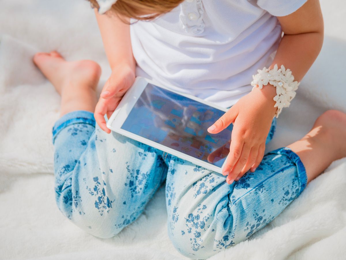 Child in denim jeans and a white top sitting on a white surface, holding a tablet.