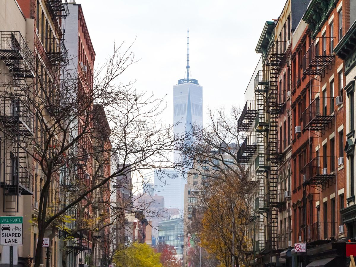 A view down a city street with brick buildings and fire escapes on both sides, and the One World Trade Center visible in the distance.